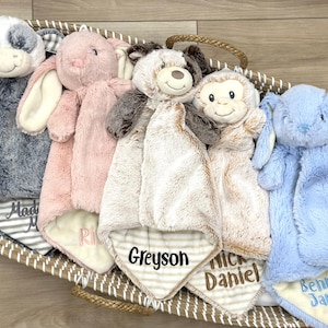 May include: A collection of plush baby security blankets in a woven basket. The blankets feature animal heads, including a cow, bunny, dog, and monkey, in various colors. Each blanket has a striped or solid-colored bottom with a name embroidered on it.