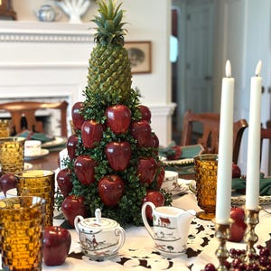 May include: A decorative centerpiece featuring a pineapple topper atop a cone-shaped arrangement of glossy red apples and green foliage. The base is surrounded by a white ceramic sugar bowl and creamer with hunting scenes, amber glasses, and two tall white taper candles.