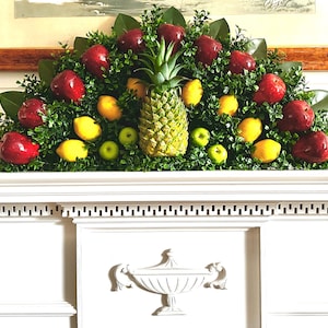 May include: A semi-circular artificial fruit arrangement featuring a central pineapple surrounded by red apples, yellow lemons, and small green apples nestled within dense green boxwood foliage and dark green magnolia leaves.