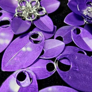 May include: A close-up of a collection of purple, metallic, leaf-shaped charms with a hole in the center of each. The charms are arranged in a random pattern on a black surface.