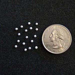 May include: A close-up of a group of small, white, round beads arranged in a circle pattern. A US quarter is included for scale.