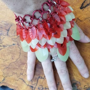 May include: A red and white chainmail bracelet with a layered design. The bracelet is made of metal rings and has a series of overlapping scales.