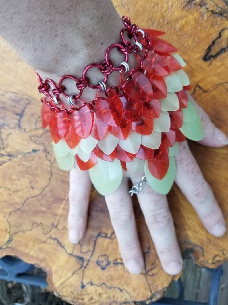 May include: A red and white chainmail bracelet with a layered design. The bracelet is made of metal rings and has a series of overlapping scales.