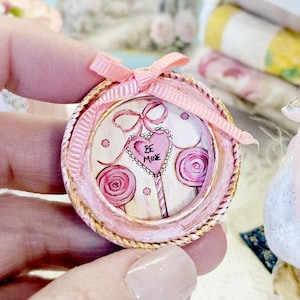 May include: A miniature round decorative frame in a distressed pink finish with a gold rope-style border. The center features a drawing of two pink lollipops and a heart-shaped candy with the text BE MINE, topped with a small pink ribbon bow.