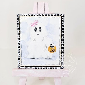 May include: A small rectangular painting on a light pink wooden easel featuring a white ghost with a pink bow on its head, holding an orange jack-o'-lantern bucket. The painting has a black and white patterned border with a light blue background.
