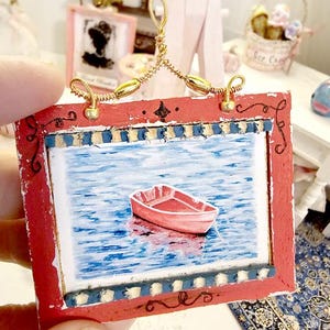 May include: Miniature red framed artwork featuring a small red boat on blue water. The frame is red with gold accents and a blue and white striped border. The artwork is held by a gold wire hanger.