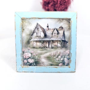 May include: A square, light blue framed artwork featuring a watercolor painting of a quaint cottage. The cottage has a gray roof, a chimney, and is surrounded by pink roses and a pathway. The frame has a distressed, vintage look with gold accents.