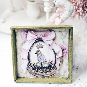 Puede incluir: Un marco cuadrado de madera con acabado verde desgastado que contiene una impresión decorativa. La imagen muestra un pájaro sentado en un nido con una corona encima, enmarcado por un gran lazo rosa. El fondo presenta un suave estampado floral.