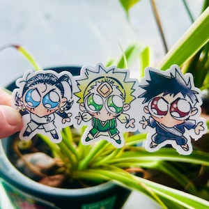 May include: Three cartoon stickers featuring anime-style characters with large eyes. The characters are in various outfits, including white, green, and navy blue. The stickers have a white border and are held in front of a green plant.