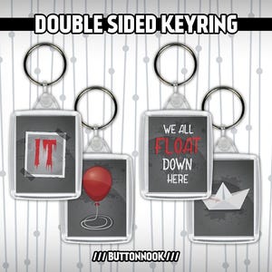 May include: Double-sided keyrings with images from the film "IT". One side features the word "IT" in red letters on a black background. The other side features a red balloon with a black string on a grey background. The second keyring features the text "We all float down here" on a black background. The other side features a paper boat on a grey background.
