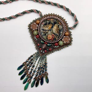 Op de afbeelding: Een kralenketting met een centrale vierkante hanger met een ronde vogelillustratie, omringd door bloemvormige en ronde kralen in groen-, rood- en bronskleuren. Een franje van druppelvormige kralen hangt aan de onderkant van de hanger.