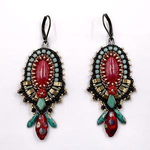 May include: Pair of ornate earrings with a red oval center stone, surrounded by turquoise and black beads. Gold and teal accents are visible, with a red and teal teardrop-shaped dangle. The earrings have a dark metal hook.