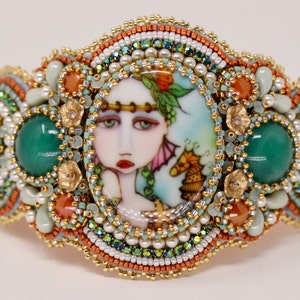 May include: A beaded bracelet with a colorful, hand-painted porcelain cameo featuring a woman's face. The bracelet is decorated with green, orange, and white beads.