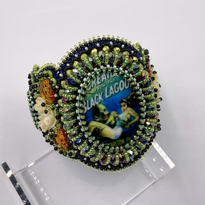 Pode incluir: Uma pulseira de punho com contas com um design circular, apresentando uma imagem de pôster de filme vintage de "Creature from the Black Lagoon". A pulseira é adornada com contas verdes, pretas e iridescentes, com detalhes em laranja e branco.
