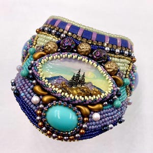 May include: A handcrafted beaded cuff bracelet in shades of blue, purple, gold, and turquoise. The bracelet features an oval centerpiece with a miniature landscape painting of mountains and trees. The design incorporates various bead sizes and textures, creating a detailed and ornate accessory.