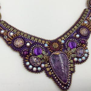 Peut inclure: Un collier artisanal en perles violettes avec une pierre centrale allongée de couleur violette. Le collier présente des perles violettes de différentes tailles, des accents irisés et des éléments métalliques dorés. Le design est complexe et orné, de forme plastron.