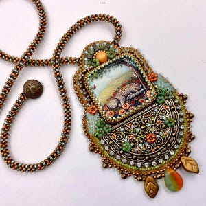 May include: Detailed pendant necklace with a miniature painting of a cat sleeping in a garden. The pendant is framed with intricate beadwork, metal filigree, and sparkling accents. The necklace has a beaded chain with a decorative clasp.