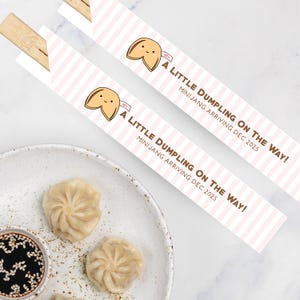 May include: Two pairs of chopsticks with a fortune cookie graphic and the text "A LITTLE DUMPLING ON THE WAY! MINIJANG ARRIVING DEC 2025". The chopsticks are placed near a plate with dumplings and a small bowl of sauce.