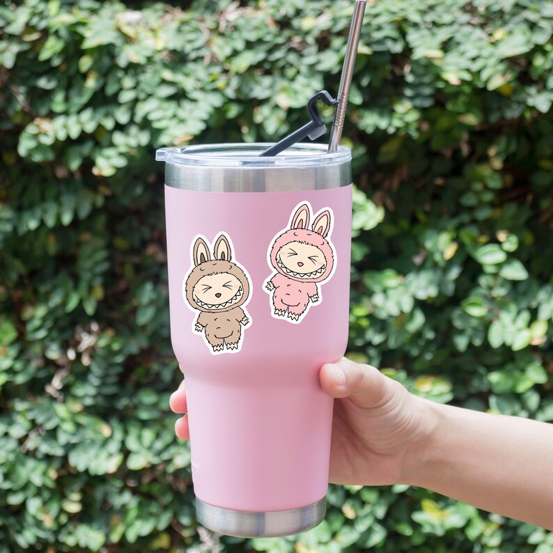 以下が含まれることがあります： ピンク色のタンブラーに銀色のストローと2つのステッカー。ステッカーは、茶色とピンクのウサギのコスチュームを着た漫画のキャラクターが特徴です。タンブラーは緑の葉の背景に対して保持されています。