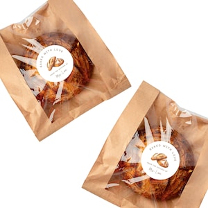 May include: Two brown paper bags with clear plastic windows, each containing a baked pastry. The pastries are round with a golden-brown crust. Each bag has a white sticker that reads "Baked with Love" and "From the Kitchen of Mrs. Lane."