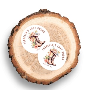 May include: Two round stickers with a watercolor design featuring a cowboy hat and boots, surrounded by flowers. The text "Isabella's Last Rodeo" and "Austin, TX" are printed on the stickers. The stickers are placed on a wooden slice.