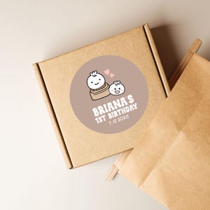 May include: A brown cardboard box and a brown paper bag are shown. A round sticker on the box features a cartoon dumpling design with the text "Briana's 1st Birthday 7.12.2025". The sticker has a light brown background with pink hearts.