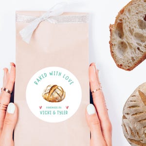 May include: A light brown paper bag tied with a white ribbon, featuring a round sticker that says "Baked with Love" and "Handmade by Vicki & Tyler." The sticker has a watercolor illustration of a loaf of bread.