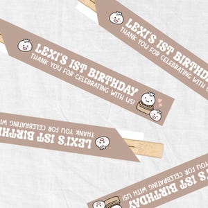 May include: Four brown and white chopsticks with a tan background. Each chopstick has a brown paper banner with a cartoon dumpling and the text "LEXI'S 1ST BIRTHDAY" and "THANK YOU FOR CELEBRATING WITH US!"