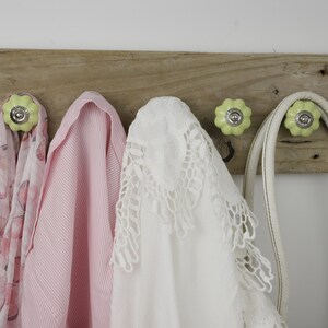 May include: A rustic wooden wall hook with four light green ceramic knobs. A pink and white scarf, a pink striped shirt, a white lace shirt, a white leather bag, and a white leather purse are hanging on the hooks.