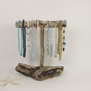 May include: A rustic wooden jewelry display stand holding various beaded necklaces in shades of blue, white, and beige. The stand is made of driftwood and has a starfish in the foreground. The necklaces are of varying lengths and bead sizes.