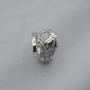 May include: A silver-toned metal ring featuring a detailed, embossed design of a bird with a long beak and folded wings. The band is smooth and polished, while the bird motif displays intricate feather textures and a high-shine finish.