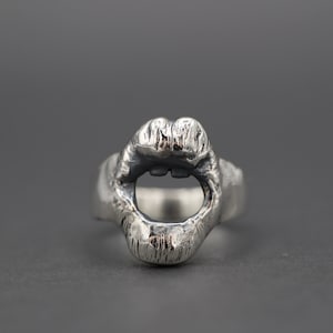 May include: A silver ring featuring a detailed design of a pair of lips. The lips are open and the ring is made from textured metal.