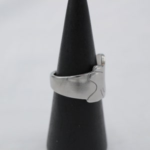 May include: A silver ring with a unique, abstract design. The ring is displayed on a black cone-shaped stand.