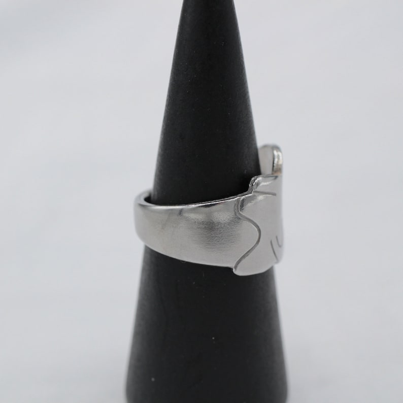 May include: A silver ring with a unique, abstract design. The ring is displayed on a black cone-shaped stand.