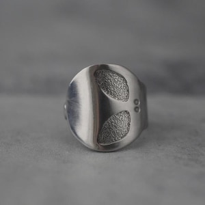 Puede incluir: Un anillo plateado con una cara ovalada. La cara presenta dos diseños texturizados en forma de almendra y dos pequeños detalles circulares. El anillo tiene un acabado liso y pulido y una banda sencilla.