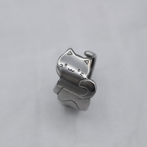 May include: A silver ring with a cat design. The cat is facing forward with its tail curled around its body. The cat has a simple, smiling face.