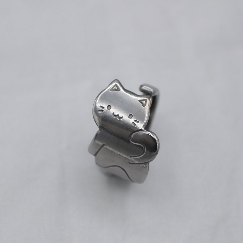 May include: A silver ring with a cat design. The cat is facing forward with its tail curled around its body. The cat has a simple, smiling face.