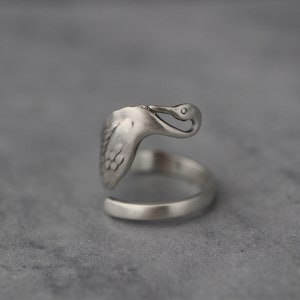 Può includere: Un anello d'argento a forma di uccello con la testa piegata verso il basso, come se stesse bevendo da una pozza d'acqua.