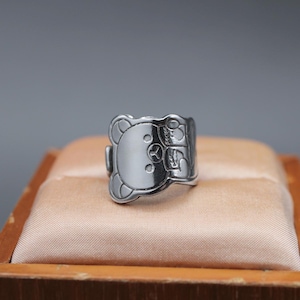 Puede incluir: Un anillo plateado con un diseño de oso. El anillo presenta una cara de oso detallada en la parte delantera, con detalles grabados en los laterales. El anillo se muestra sobre una superficie de color melocotón.