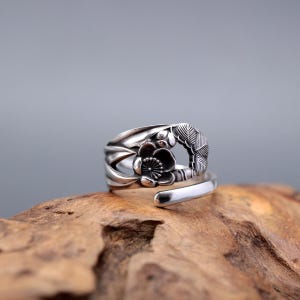 Op de afbeelding: Een zilverkleurige ring met een bloemmotief. De ring heeft een gedetailleerd bloem- en bladmotief. De ring ligt op een houten oppervlak.