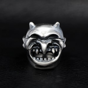 May include: A silver ring with a detailed design of a grinning demon face with horns and sharp teeth.