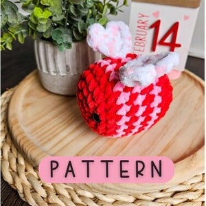 Peut inclure: Un jouet amigurumi en crochet en forme d'abeille avec des ailes roses et rouges et un corps rouge et rose. Le jouet est posé sur une surface en bois avec un calendrier qui indique "février 14" en arrière-plan. Le texte "PATTERN" est écrit en rose sur fond blanc.