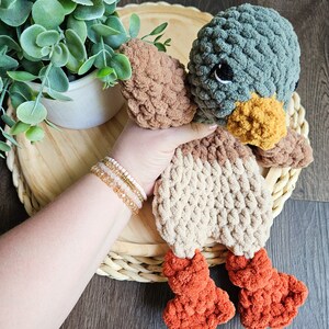 Puede incluir: Un juguete de peluche de pato tejido a crochet con hilo marr&oacute;n, beige y naranja. El pato tiene una cabeza verde con ojos negros y un pico amarillo.