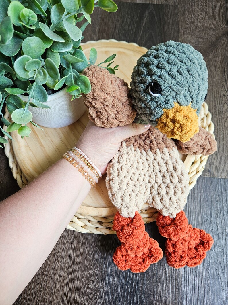 Puede incluir: Un juguete de peluche de pato tejido a crochet con hilo marr&oacute;n, beige y naranja. El pato tiene una cabeza verde con ojos negros y un pico amarillo.