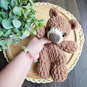 May include: A handmade brown teddy bear with a white muzzle and dark eyes. The bear is made of a soft, textured yarn and is sitting on a wooden tray. A floral bracelet is visible in the image.