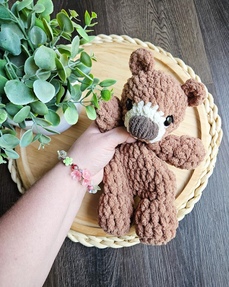 May include: A handmade brown teddy bear with a white muzzle and dark eyes. The bear is made of a soft, textured yarn and is sitting on a wooden tray. A floral bracelet is visible in the image.