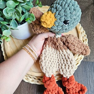 Puede incluir: Un juguete de peluche de pato tejido a crochet con cabeza turquesa, cuerpo marr&oacute;n y beige y patas naranjas. El pato est&aacute; sentado en una cesta tejida.