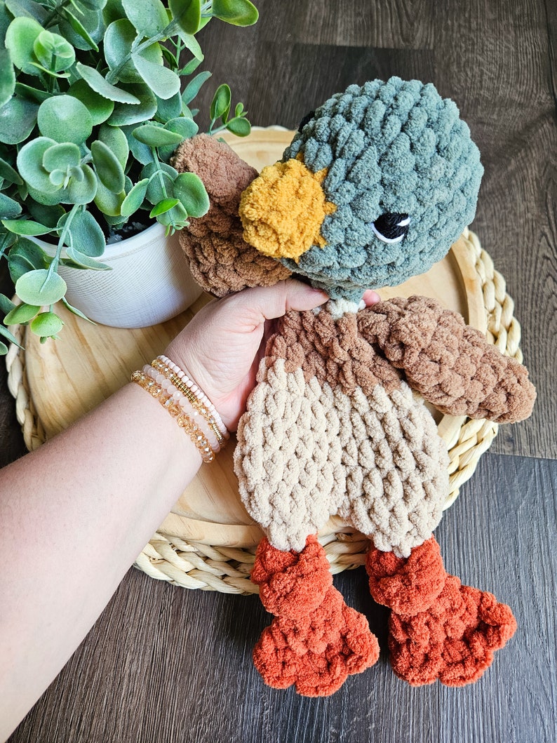 Puede incluir: Un juguete de peluche de pato tejido a crochet con cabeza turquesa, cuerpo marr&oacute;n y beige y patas naranjas. El pato est&aacute; sentado en una cesta tejida.