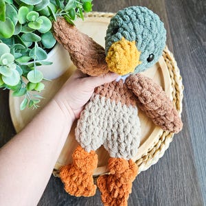 Puede incluir: Un juguete de peluche de ganchillo en forma de pato con cabeza turquesa, cuerpo marr&oacute;n y patas naranjas. El pato est&aacute; sentado en una bandeja de madera con un patr&oacute;n tejido.