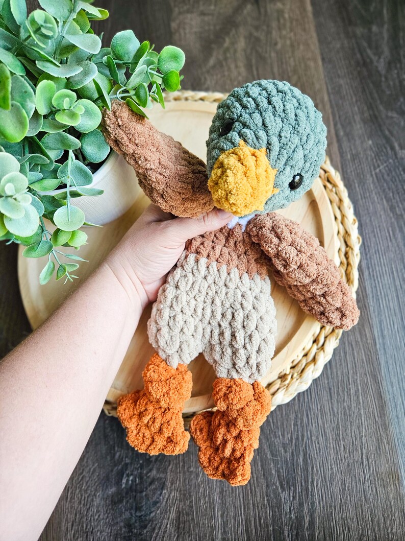 Puede incluir: Un juguete de peluche de ganchillo en forma de pato con cabeza turquesa, cuerpo marr&oacute;n y patas naranjas. El pato est&aacute; sentado en una bandeja de madera con un patr&oacute;n tejido.